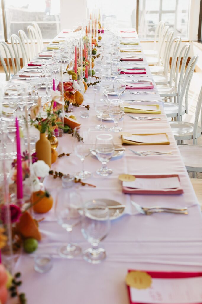 A long dining table is elegantly set with white linens, colorful napkins, candles, and floral centerpieces. Wine glasses and cutlery are neatly arranged at each place setting.