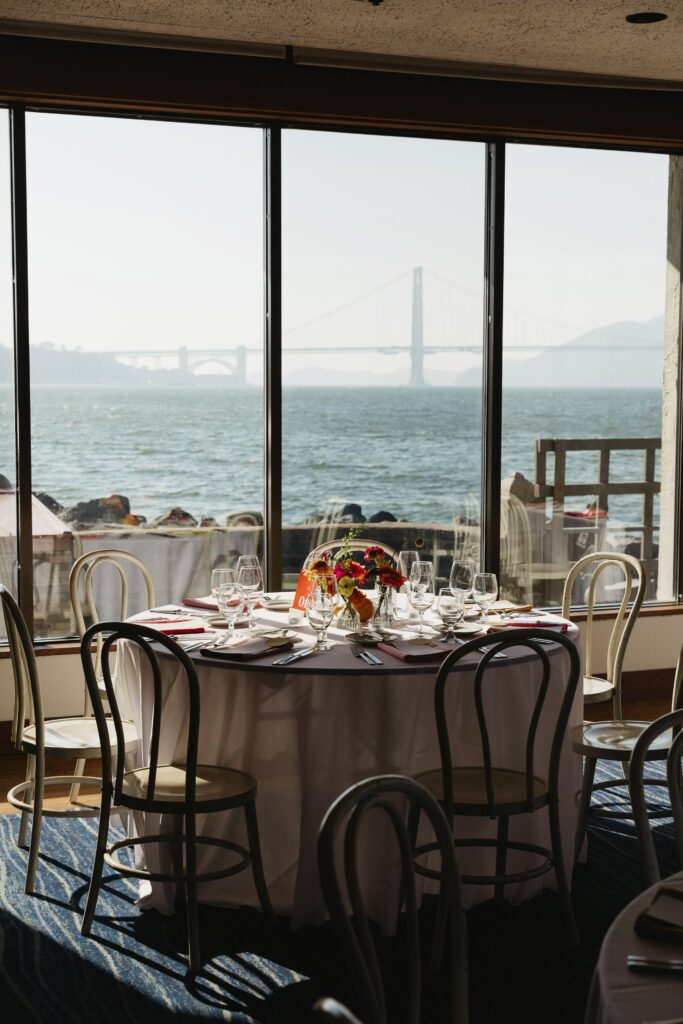 A round dining table set for six by a window with a view of a large bridge over water in the background.