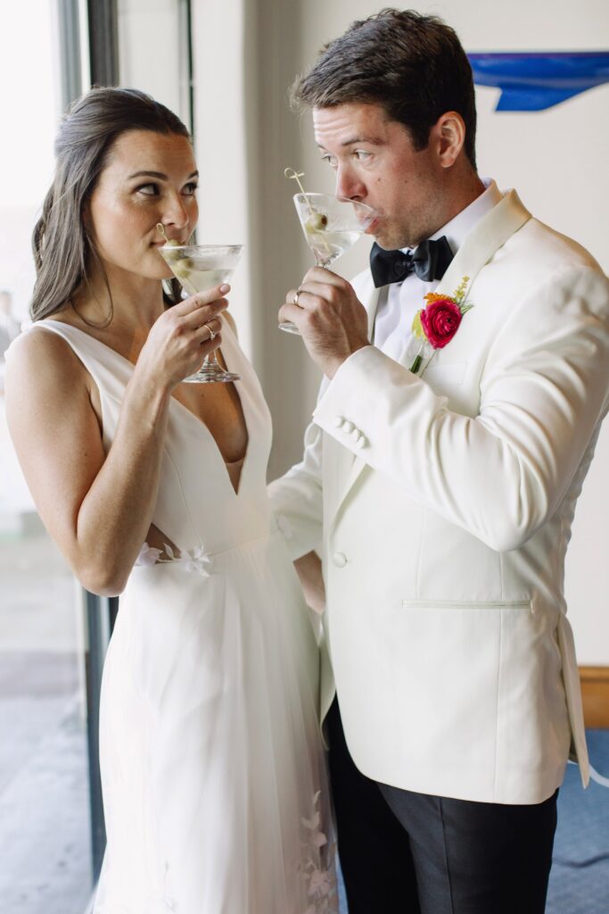 A couple in formal attire, both wearing white, enjoy cocktails with a green olive garnish. The woman looks towards the man as they both raise their glasses.