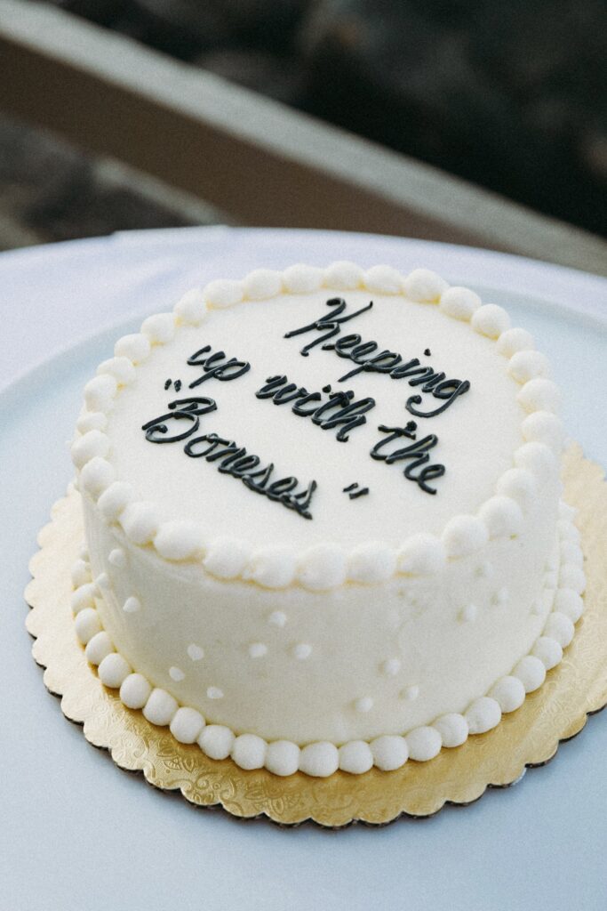 Round white cake with piped icing words reading "Keeping up with the Boness" on top, surrounded by a decorative white border, on a gold and white base.