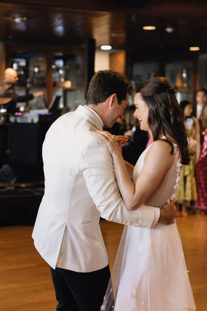 A couple in formal attire dances closely in a warmly lit room with people visible in the background.