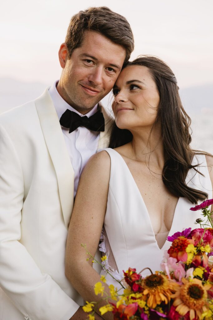 A couple in wedding attire, with the man in a white suit and bow tie, and the woman in a white dress holding a colorful bouquet, stand close with a serene backdrop.