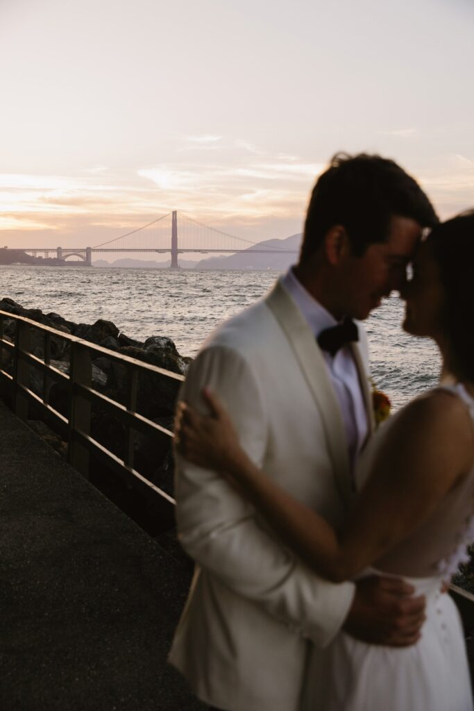 A couple embraces near a railing with a view of the Golden Gate Bridge in the background during sunset.