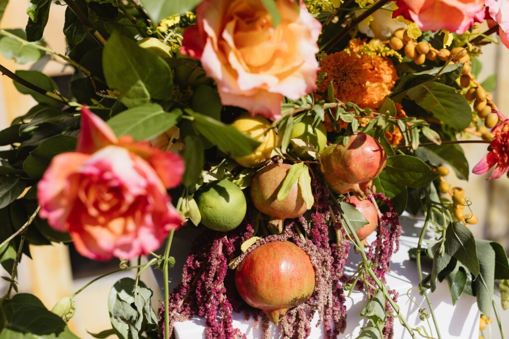 A vibrant floral arrangement featuring roses, marigolds, greenery, pomegranates, and various fruits.