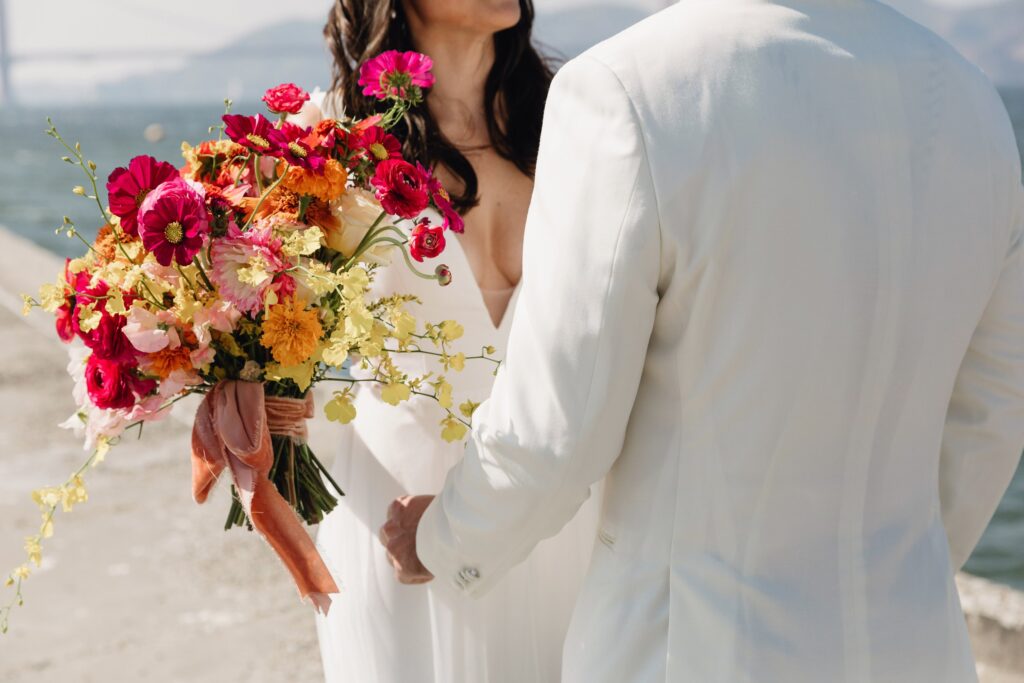 Bride and groom in white attire stand by the water, with the bride holding a colorful bouquet of flowers.
