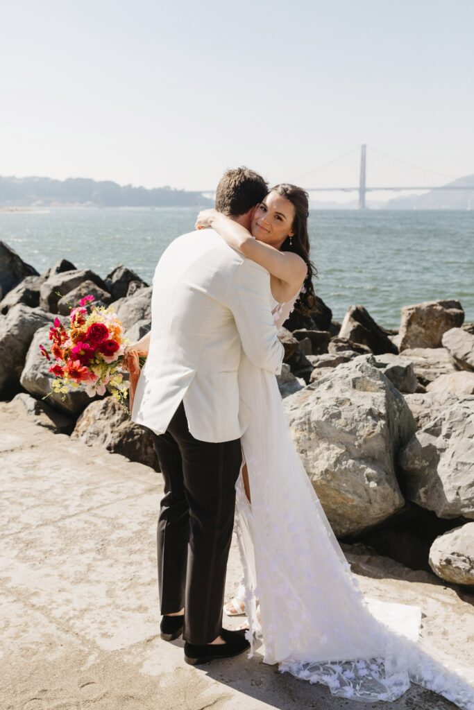 A couple in wedding attire embrace near a rocky shore with a bay and bridge in the background. The bride holds a colorful bouquet.