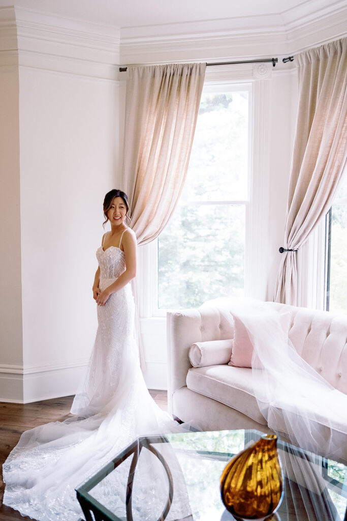 The bride in a lace wedding dress stands by a window in the bright room, reminiscent of the elegant ambiance of Park Winters Weddings, with a sofa and coffee table completing the serene scene.
