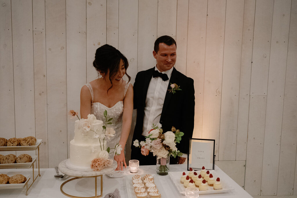 A bride and groom stand by a table with a cake, flowers, and desserts, basking in the charm of Park Winters Weddings, in front of a white wooden wall.