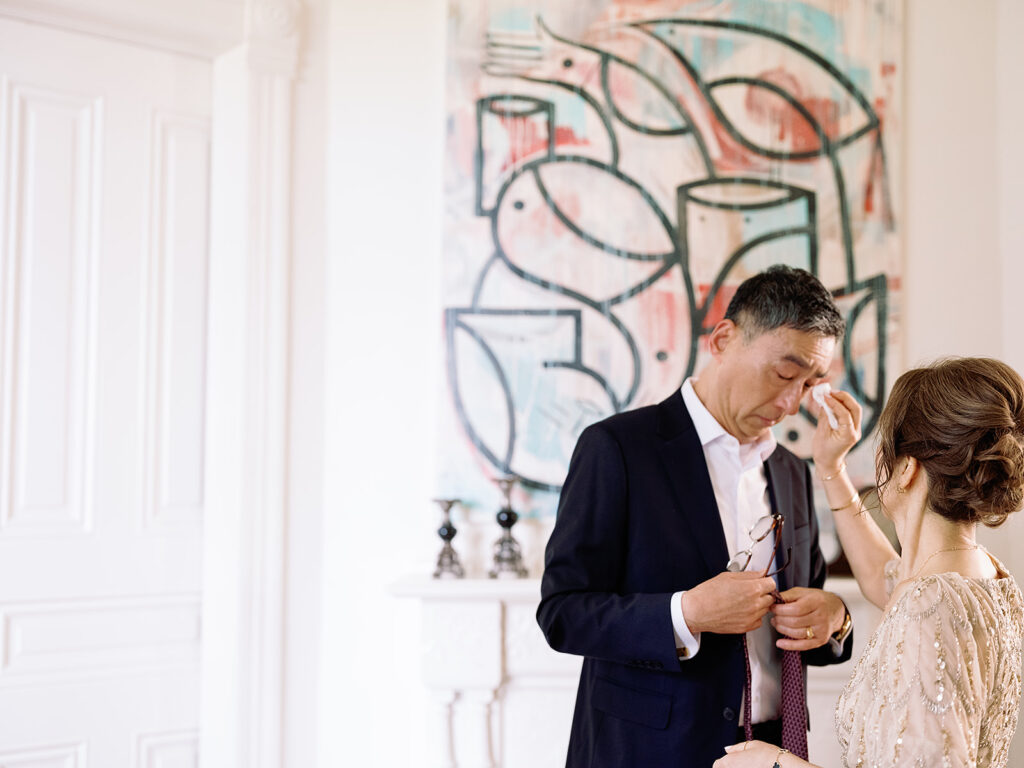 A man in a suit and a woman in a patterned dress stand before abstract art, reminiscent of Park Winters Weddings, in a room with white walls and an elegant mantle.