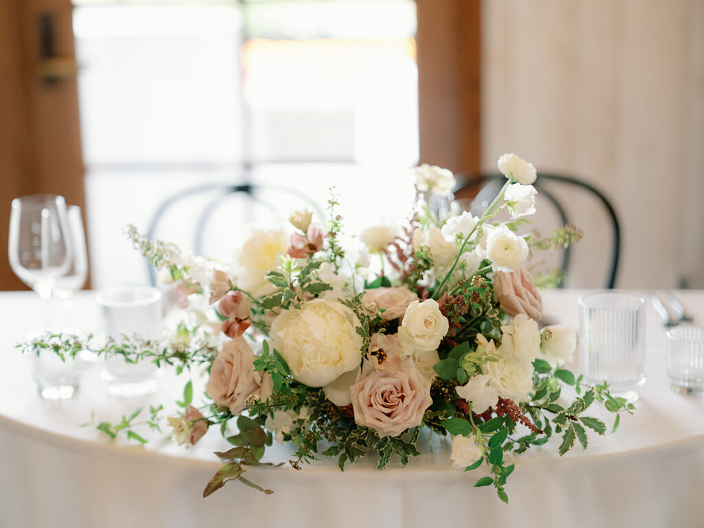 A floral centerpiece with white, cream, and pale pink flowers graces a round table at Park Winters Weddings, surrounded by empty wine glasses and small clear containers.