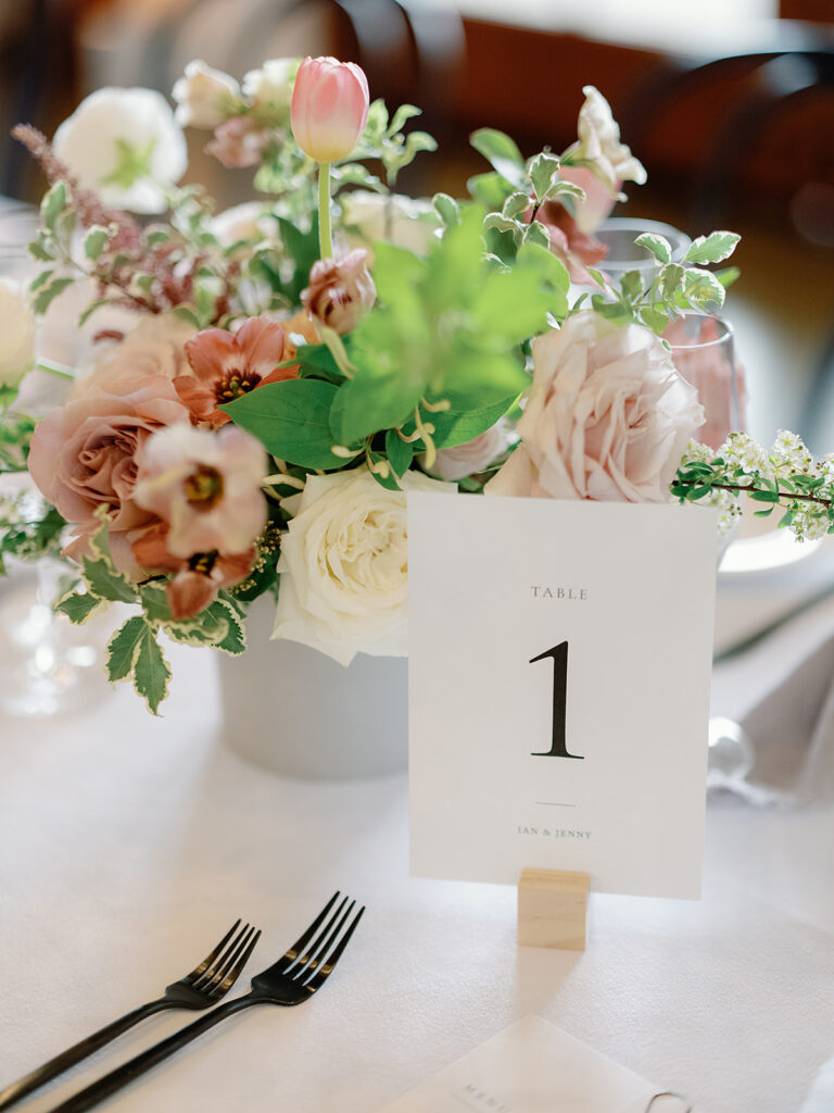 Floral centerpiece with pink and white flowers gracing a Park Winters Weddings table setting, featuring a "Table 1" sign amidst decorative greenery and place settings with forks visible.