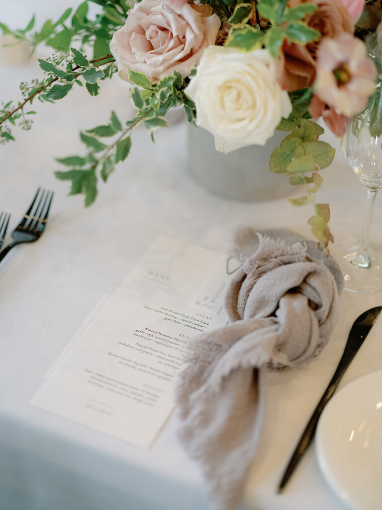 A floral arrangement with pink and white roses graces the table at Park Winters Weddings, accompanied by a menu, a beige napkin, forks, a knife, a plate, and a wine glass.