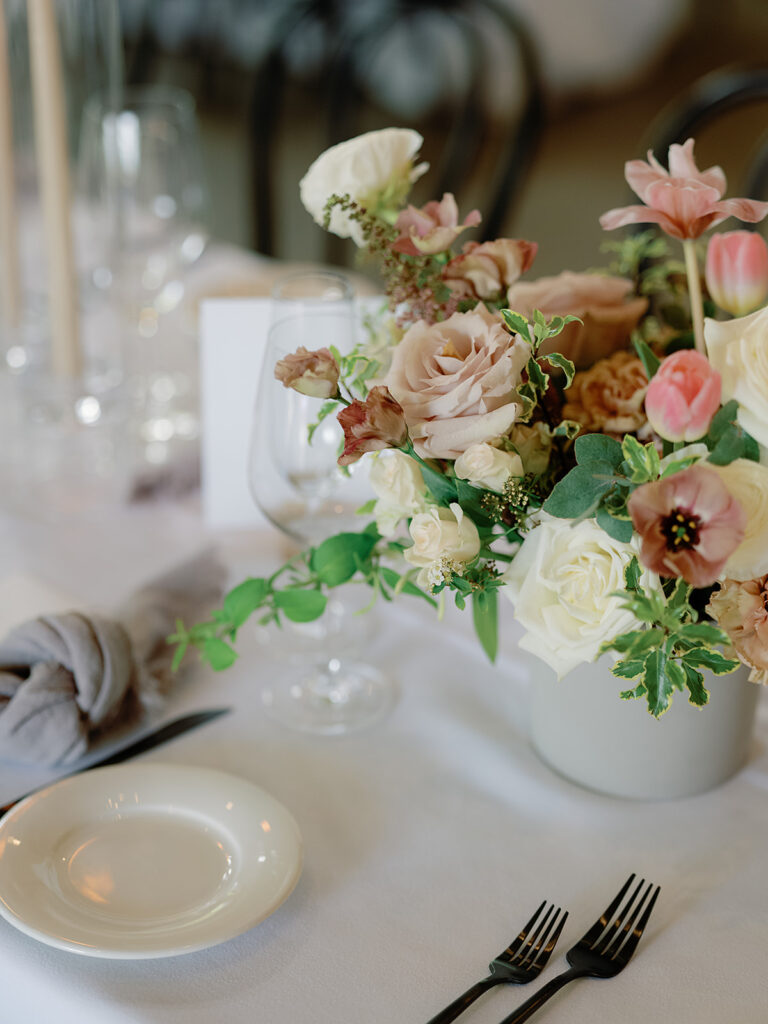 A table setting perfect for Park Winters Weddings, featuring a floral centerpiece with pastel roses and tulips, surrounded by elegant glassware, a white plate, and black cutlery on a crisp white tablecloth.
