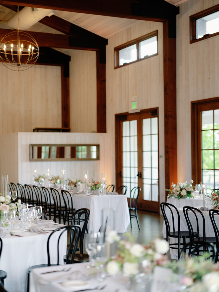 Experience the elegance of Park Winters Weddings with a charming dining setup featuring round tables adorned with white tablecloths, black chairs, and floral centerpieces, all lit by a stunning chandelier in a warm wooden venue.