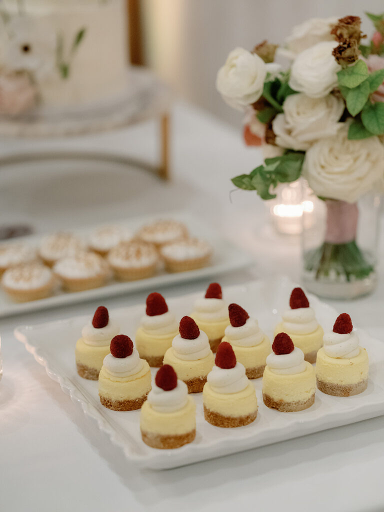 A plate of small cheesecake bites topped with raspberries sits in the foreground, echoing the elegance of Park Winters Weddings, with a vase of white flowers and a tray of cupcakes beautifully arranged in the background.