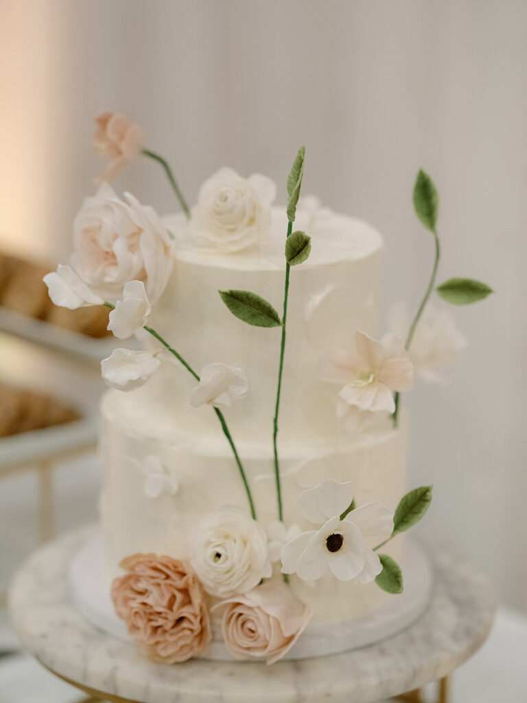 A two-tiered white cake, perfect for Park Winters Weddings, adorned with delicate pink and white flowers and green leaves, rests elegantly on a marble table.