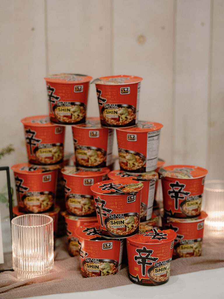 Stacked red containers of Shin Ramyun noodle soup against a light wood background, with candles reminiscent of a Park Winters wedding setting on the sides.