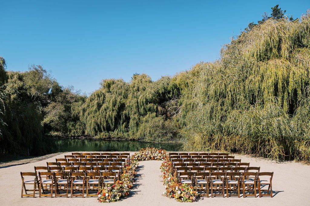 Outdoor wedding setup with rows of wooden chairs arranged along a sandy aisle, decorated with floral arrangements. Surrounded by lush greenery and a pond in the background under a clear blue sky.