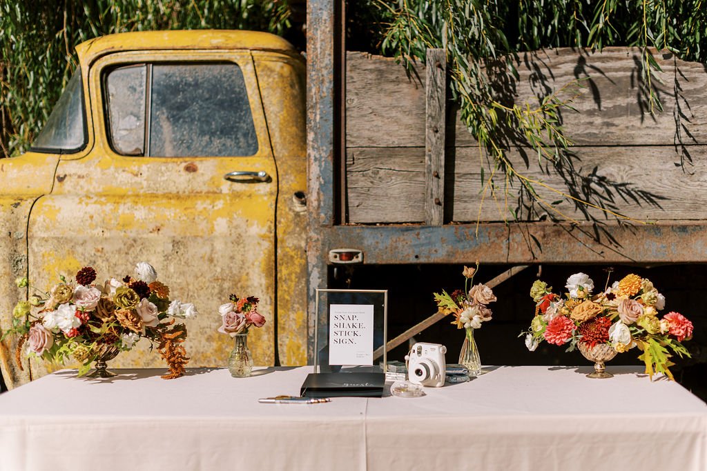 A vintage truck serves as a backdrop for a table with floral arrangements, a sign, a camera, and a guestbook on a white tablecloth.