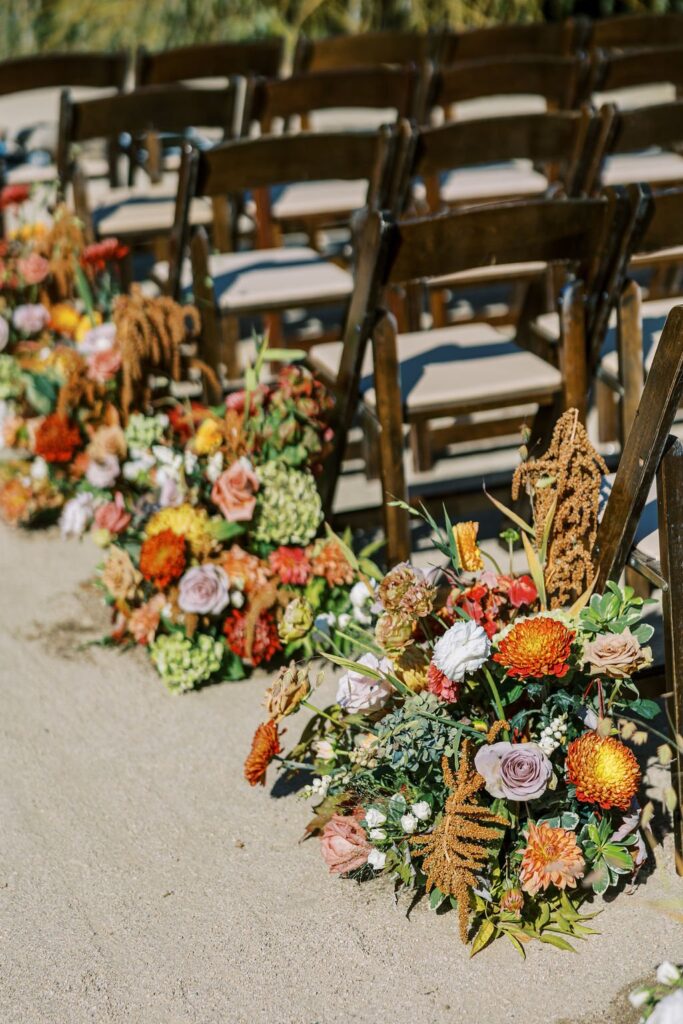 Wooden chairs are lined up in rows on a sandy surface, decorated with colorful floral arrangements including orange, yellow, and pink flowers.