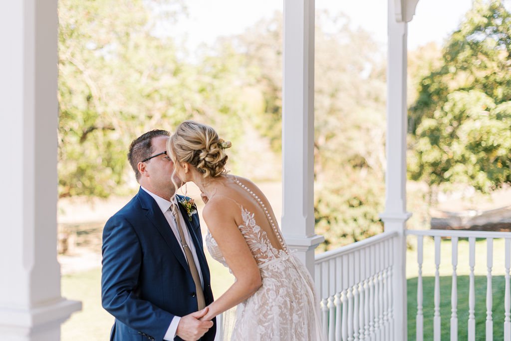 A couple kissing on a porch, with the woman wearing a lace wedding dress and the man wearing a blue suit. Trees and grass are visible in the background.