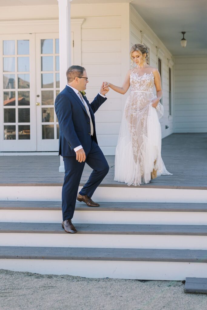 A man in a suit helps a woman in a lace wedding dress down a set of steps outside a white building.