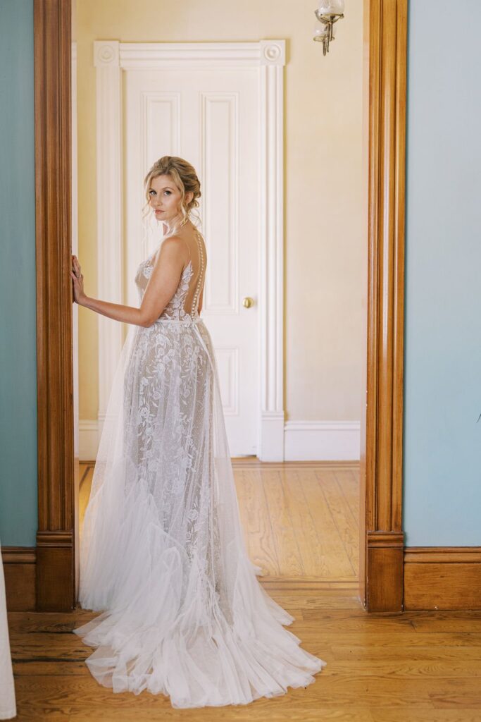 A woman in a white lace wedding dress stands in a doorway, looking back over her shoulder. She is in a room with wooden floors and a white door.