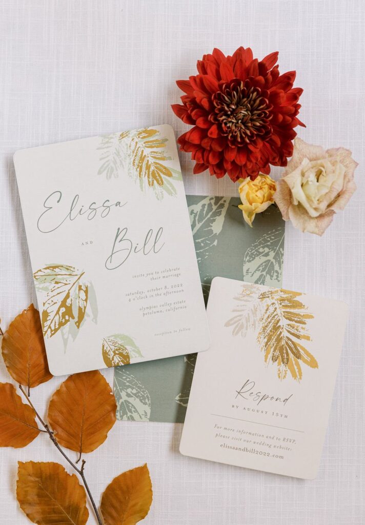 Wedding invitation suite with autumn leaf designs, featuring a large red dahlia, pale rose, and orange leaves. Text reads "Elissa and Bill" with RSVP details.