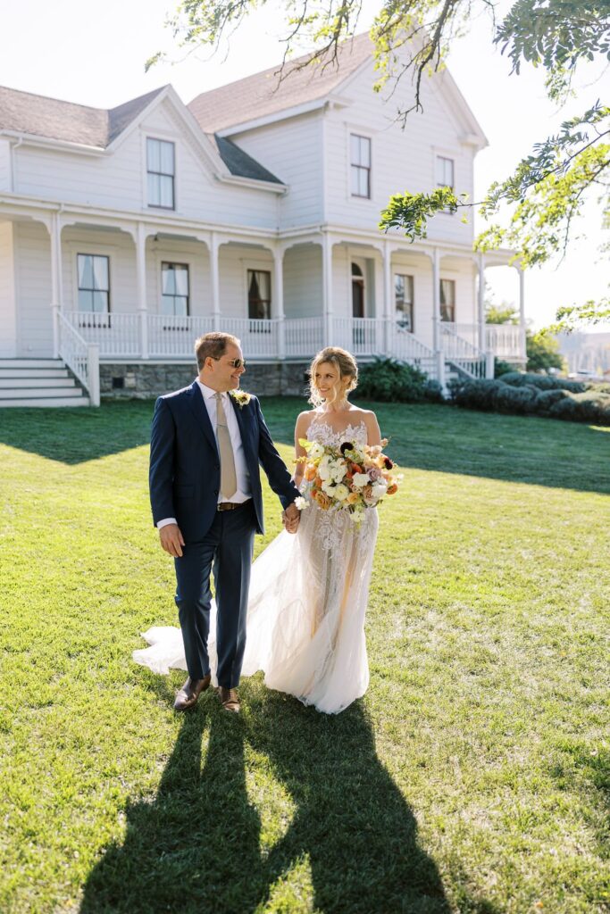 A couple walks hand in hand on a grassy lawn in front of a white house. The woman holds a bouquet of flowers.