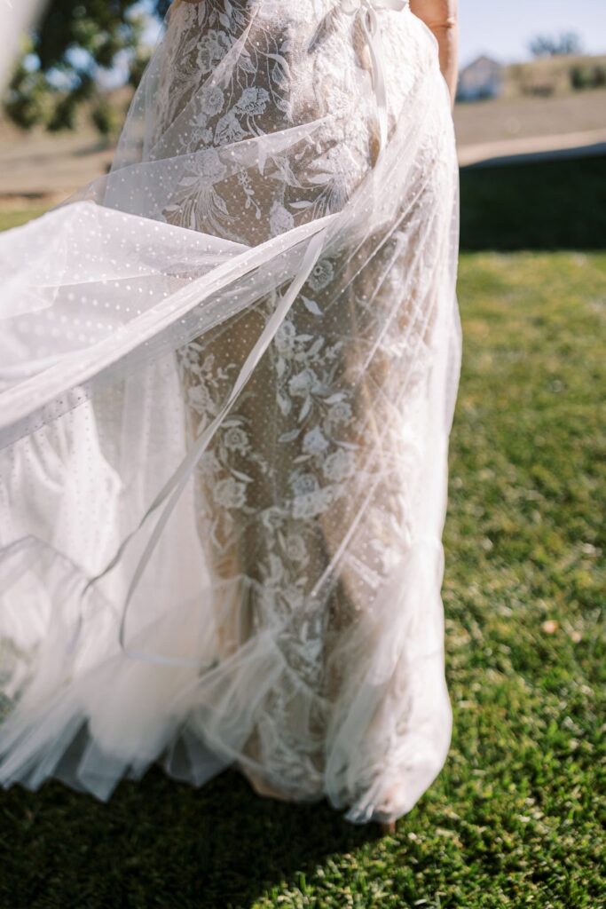 A person wearing a flowing, translucent lace dress stands on grass, with the fabric gently blowing in the breeze.