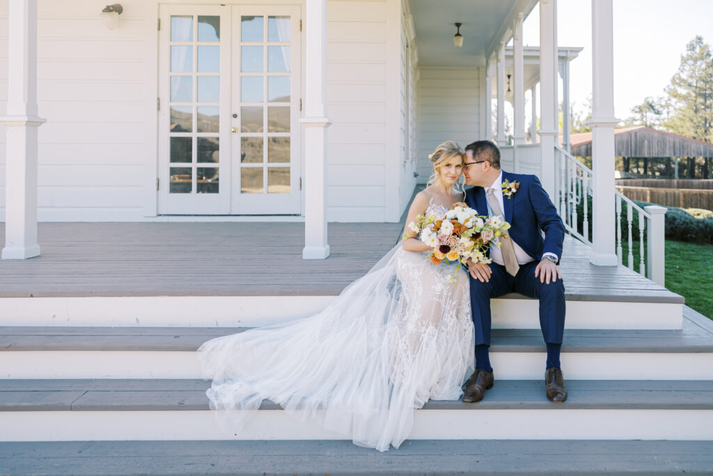 A bride in a white gown and a groom in a navy suit sit on porch steps, holding a bouquet and smiling, with a white house and glass doors in the background.