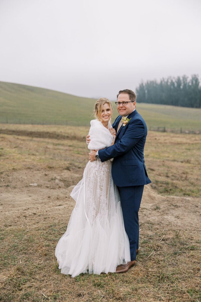 A bride and groom stand in an outdoor field. She wears a white gown and shawl; he wears a blue suit. They smile while embracing, with a grassy hill and overcast sky in the background.
