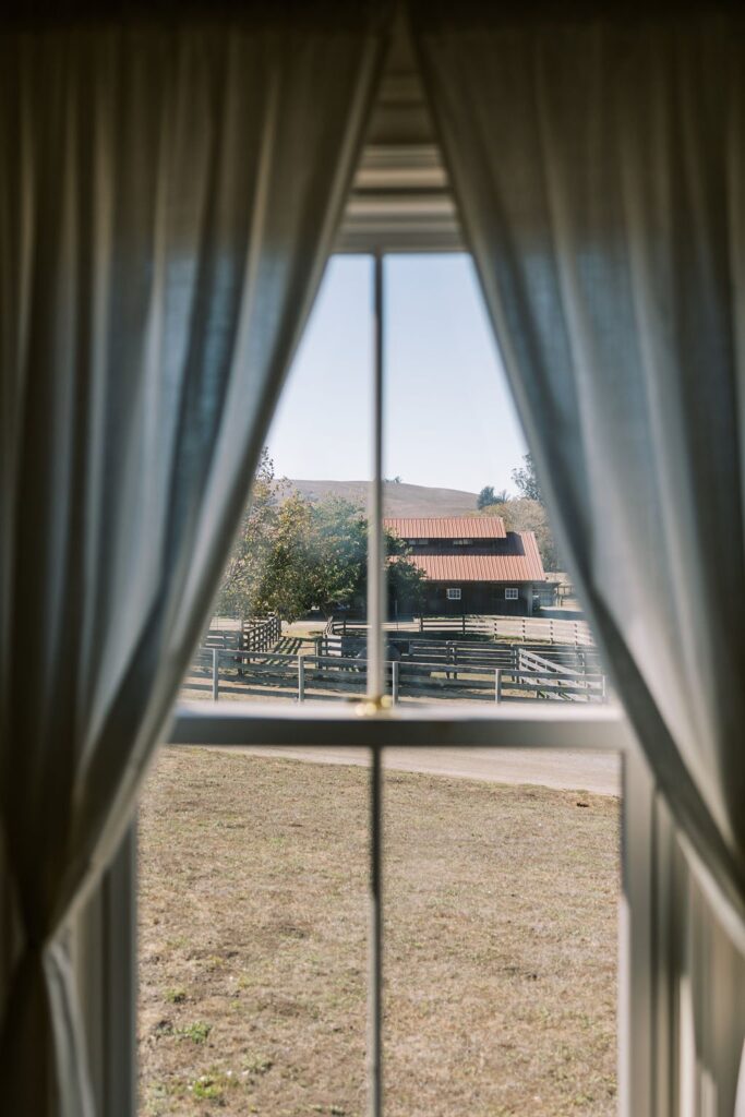 View of a rustic house with a red roof and wooden fence, seen through a window with sheer white curtains.