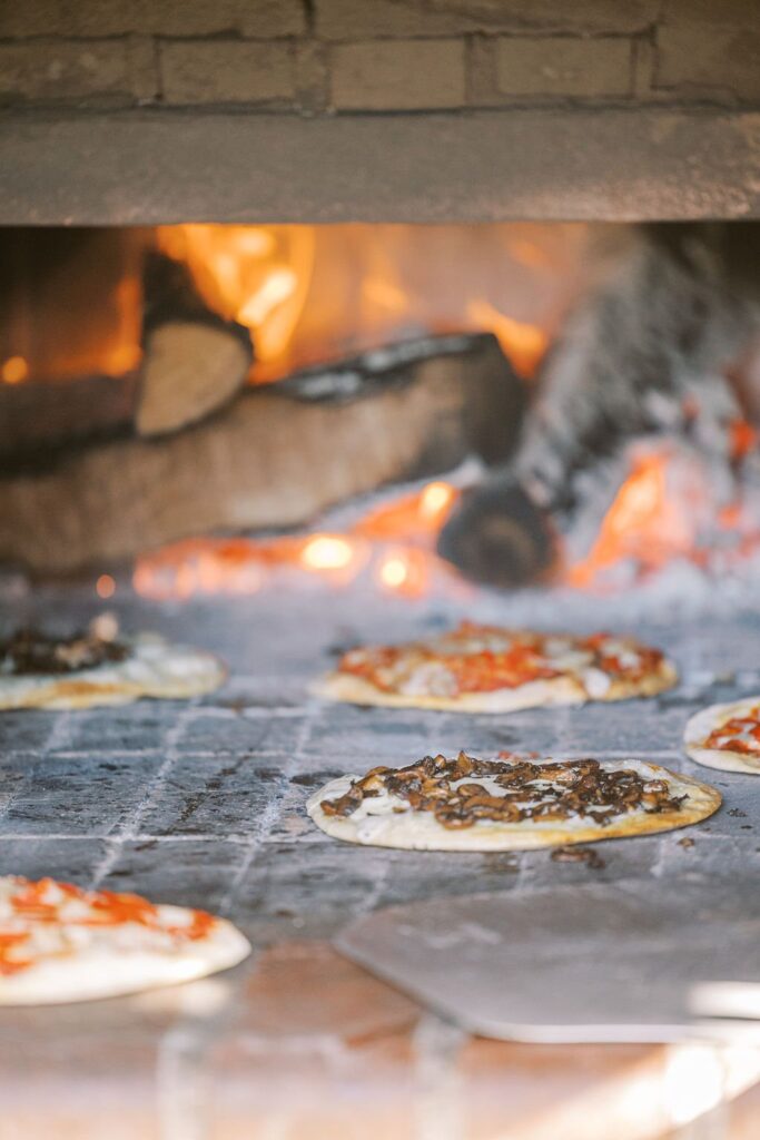 Pizzas baking in a wood-fired oven with visible flames and stacked logs in the background.