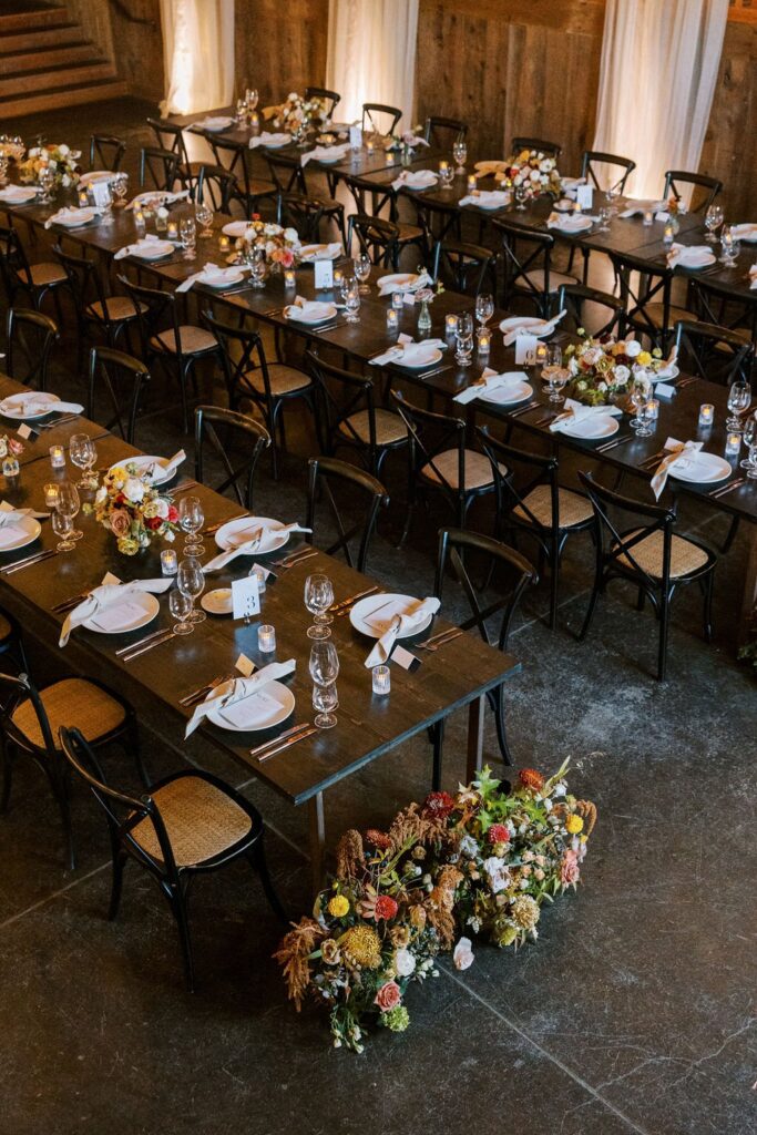 A long wooden table set with white plates, cutlery, and glassware. Chairs surround the table, and floral arrangements are placed at intervals, including one large display on the floor.