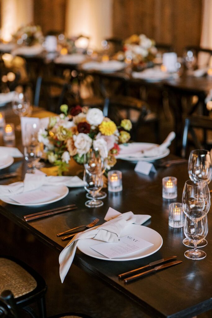 Elegant dinner table with white plates, folded napkins, wine glasses, candles, and a floral centerpiece.