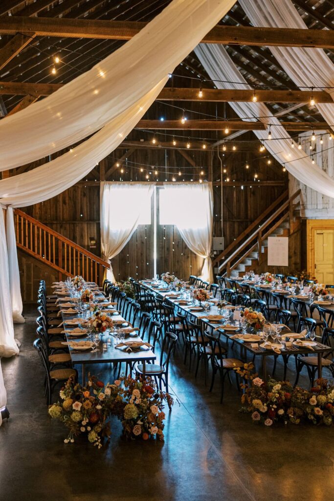 A rustic event hall with wooden beams and draped fabric, featuring long banquet tables set with tableware and floral centerpieces, illuminated by string lights.