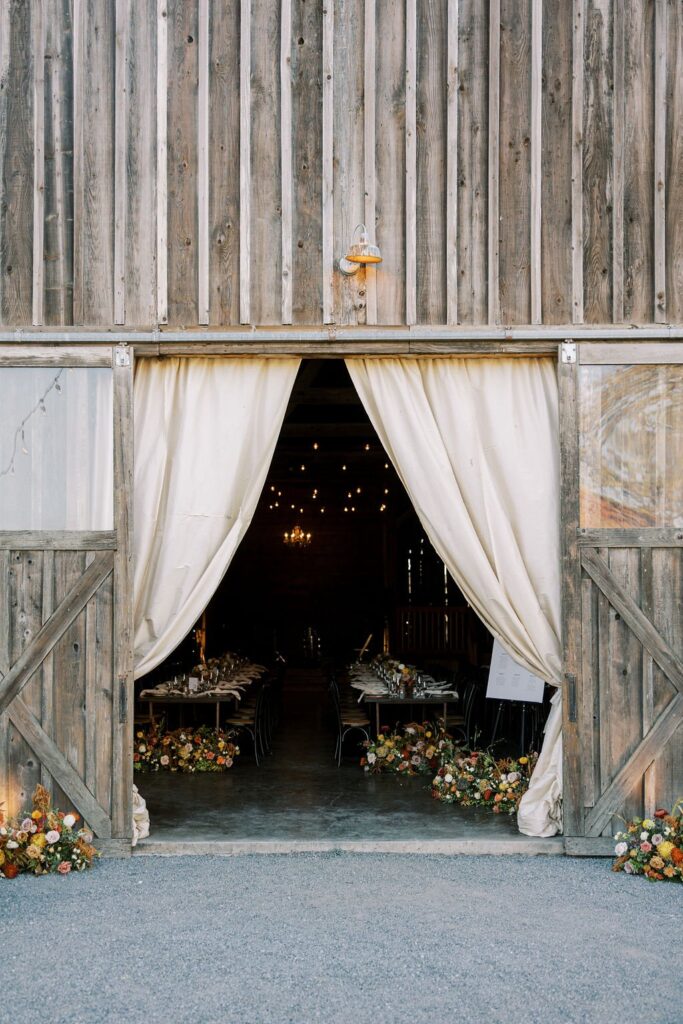 Entrance to a rustic barn with open wooden doors and white curtains, revealing a table set for an event. String lights and small floral arrangements line the entrance.