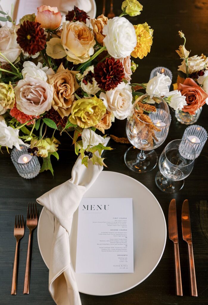 Elegant table setting with a menu card on a plate, surrounded by a floral centerpiece, wine glasses, and utensils. Roses and candles enhance the decor on a dark wooden table.