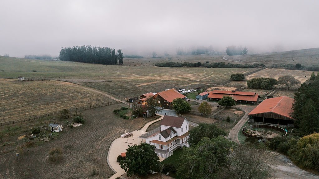 Aerial view of a farm with scattered buildings, fields, and trees under a cloudy sky.
