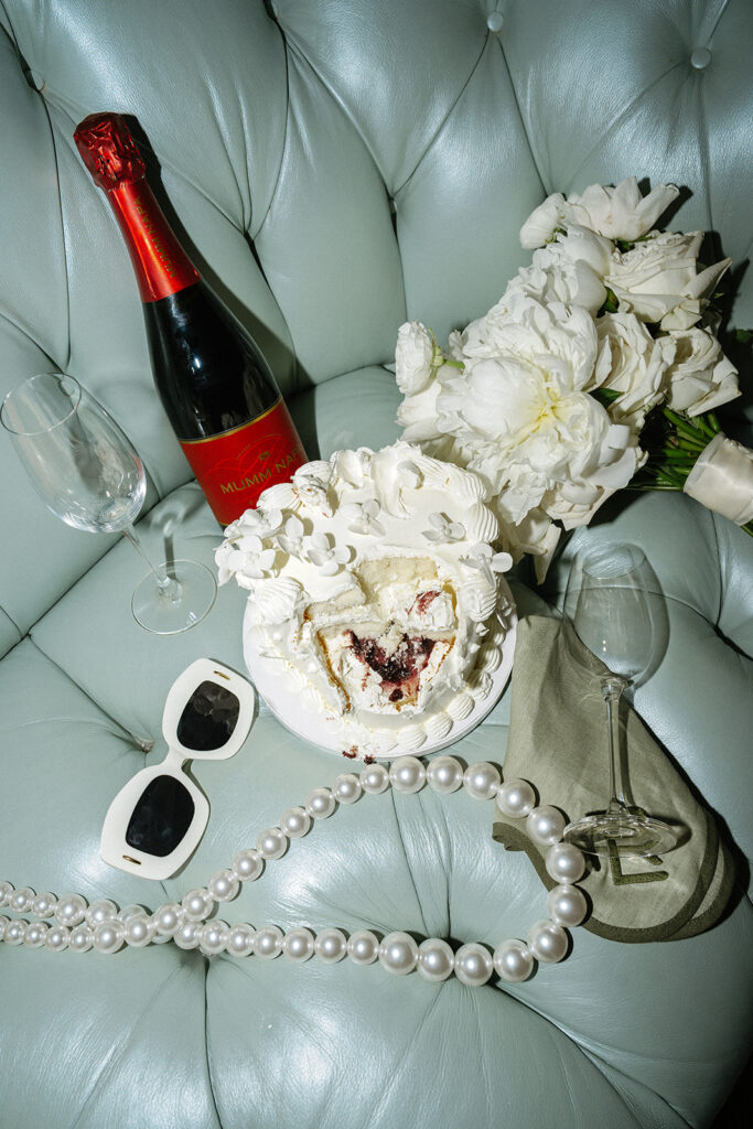 A cake slice, champagne bottle, two glasses, white flowers, sunglasses, and pearl necklace arranged on a light blue leather surface.