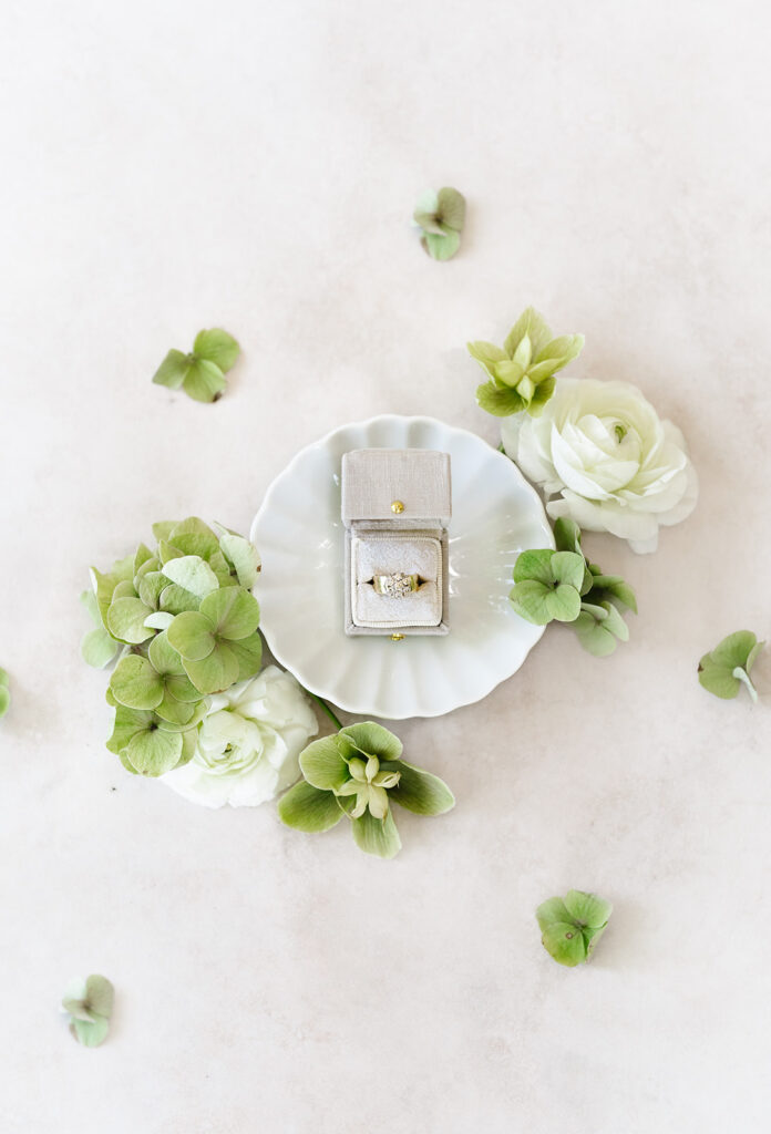 A gold ring with a diamond in a gray box on a small white dish, surrounded by green leaves and white flowers.