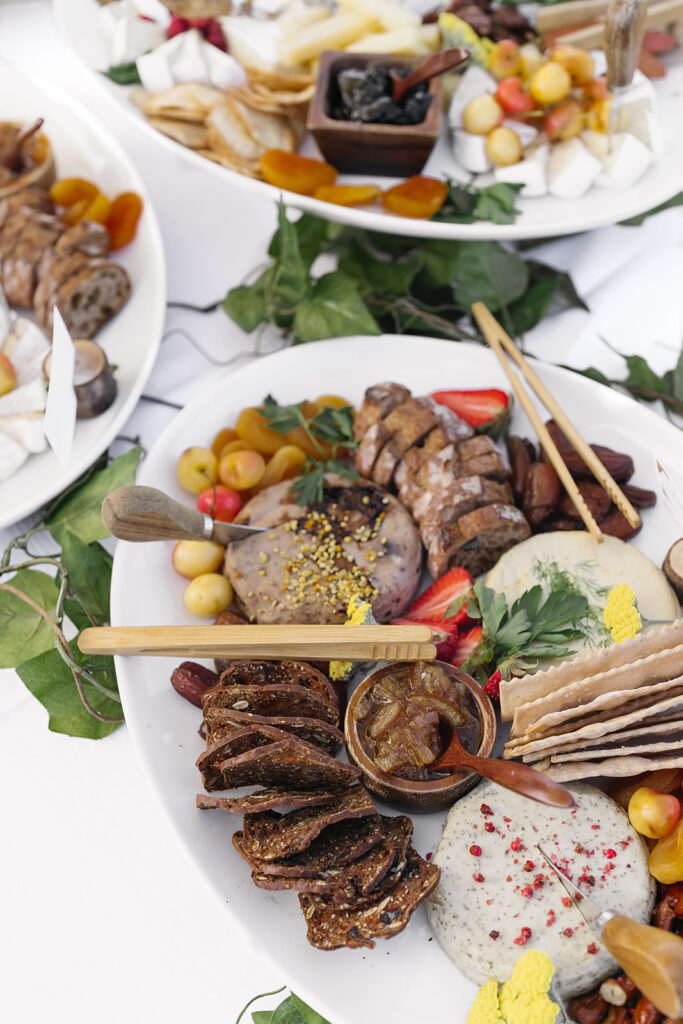 A charcuterie platter with various meats, cheeses, crackers, fruit, and dips, garnished with herbs and flowers on a white table.