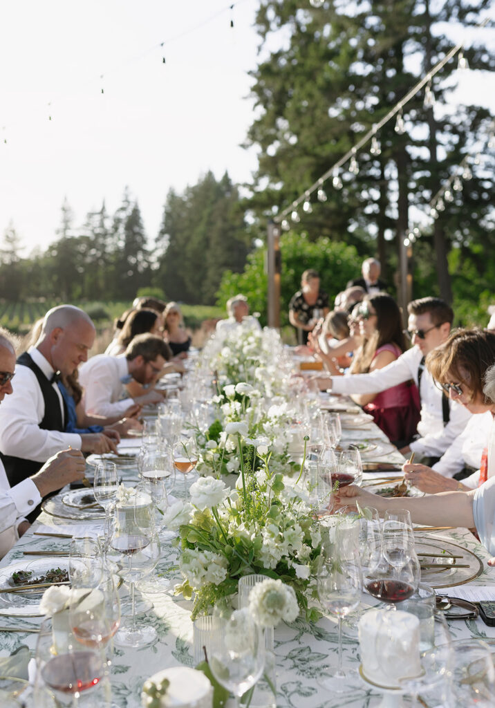 Outdoor dinner gathering with people seated at a long table decorated with white flowers, under string lights, and surrounded by trees.