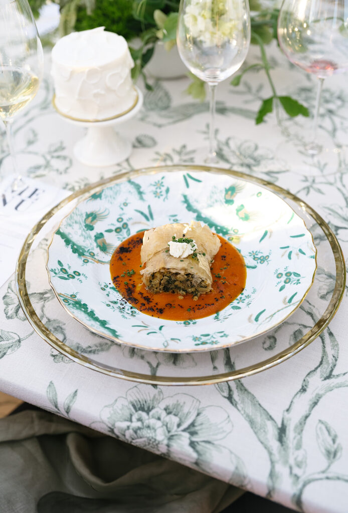 A gourmet dish of savory strudel on a patterned plate, garnished with a sauce and herbs, set on an elegant floral tablecloth with wine glasses and a small cake in the background.