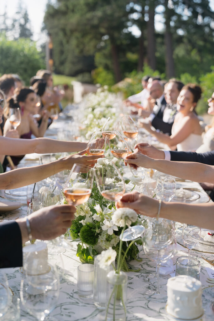 Guests clink glasses at a long, outdoor dining table adorned with flowers, celebrating in a lush garden setting.