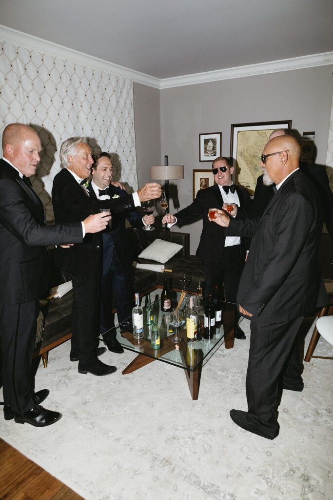 A group of men in suits stand in a living room holding drinks, toasting around a glass coffee table with bottles.