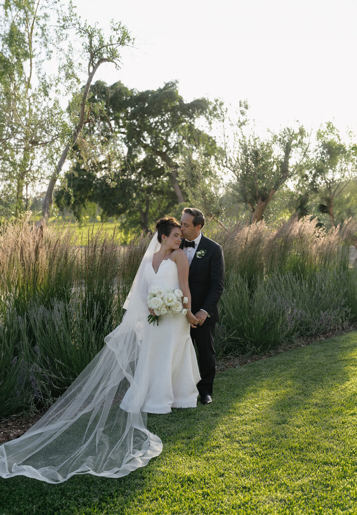 A bride and groom stand on a grassy lawn, with the groom kissing the bride's forehead. She holds a bouquet and wears a long veil. Trees and tall grasses are in the background.