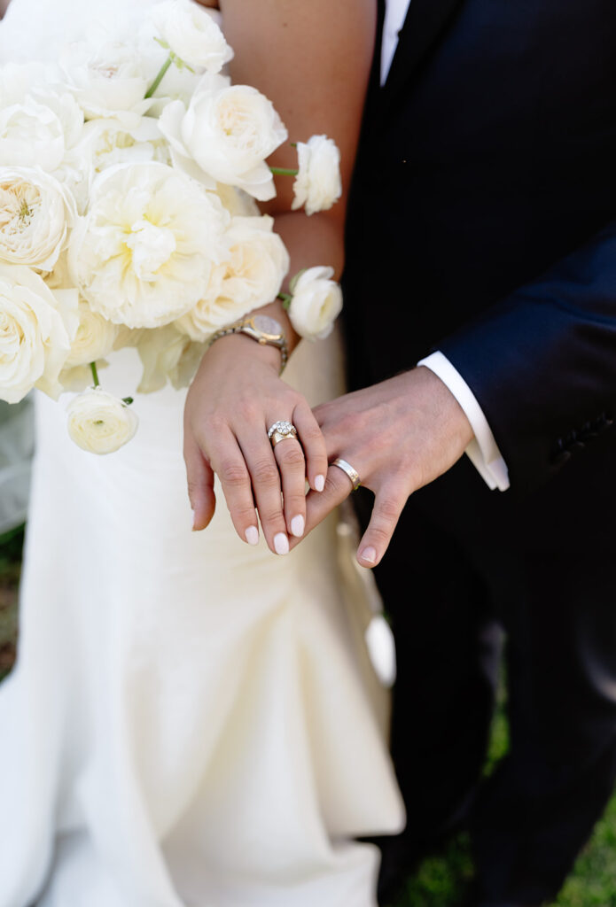 Bride and groom displaying wedding rings; bride holds a bouquet of white flowers.