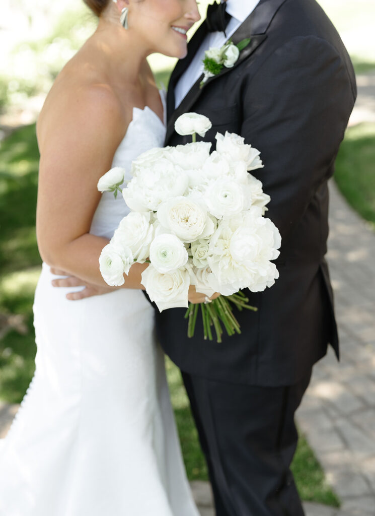 A bride in a white dress holds a bouquet of white flowers while standing next to a groom in a black tuxedo. They are outside on a paved path with grass in the background.
